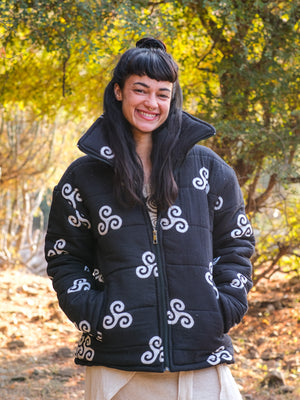 A woman with long dark hair and bangs smiles outdoors, wearing a stylish black Niruha Jacket adorned with white spiral designs inspired by Indian fashion. Trees and sunlight create a vibrant background.