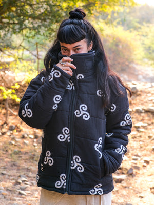 A person with long dark hair styled in a topknot stands outdoors, wearing the Niruha Jacket—a black puffer featuring white swirl designs—and partly covers their face with its high collar. Sunlight filters through the trees behind them, evoking a subtle connection to Indian fashion.