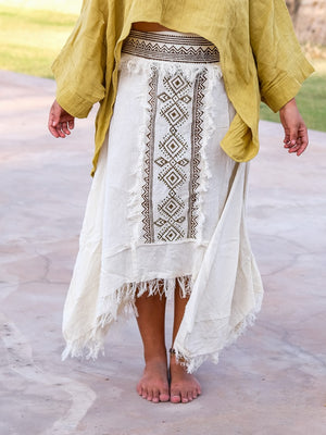 Lower-body view of the white Nirvighna Skirt on stone paving with a mustard shawl falling over the hips, showing the brown hand block printed tribal centre panel with fringed edging and the raw-fringed layered asymmetrical hem of this cotton maxi skirt.