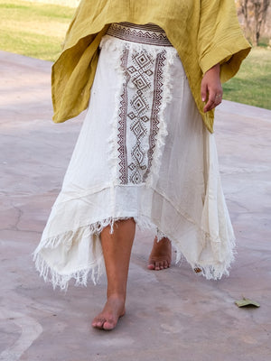 Model walking barefoot on stone paving in the white Nirvighna Skirt with a mustard shawl over her shoulders, showing the brown hand block printed tribal centre panel with fringed edging and the raw-fringed layered asymmetrical cotton hem.