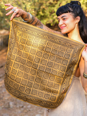 A smiling person with tattoos holds up the green Presh Bandana, a square block-print accessory featuring a repeating black spiral pattern and decorative border, while standing outdoors in natural sunlight.