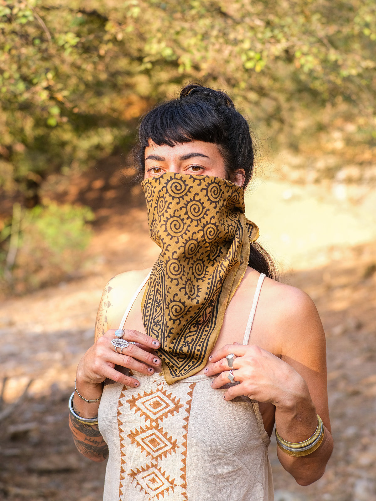 Wearing the Presh Bandana over their nose and mouth, a person stands outdoors dressed in a sleeveless top with geometric designs, accessorized with rings and bracelets against a natural, sunlit backdrop. The Presh Bandana is a versatile accessory that adds a touch of bohemian flair to this effortlessly stylish look.