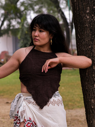 A woman with long dark hair stands near a tree, wearing a boho gypsy outfit. Her halter neck crop top, the Shanti Crop Top in dark brown, pairs with a patterned white wrap skirt. She gazes thoughtfully to the side with one arm resting on the tree trunk amidst lush greenery.