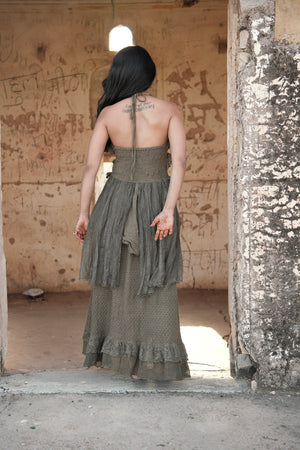 A person seen from behind wears the olive green Rust Ember Nomad Maxi Dress, a floor-length, bohemian halter-neck style with a textured bodice and ruffled hem, standing in the doorway of a weathered stone building.