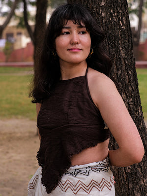 A woman with long dark hair stands by a tree in a park, gazing to her right. She is wearing a brown Shanti Crop Top and a boho gypsy patterned skirt. The background features grass, trees, and blurred structures.