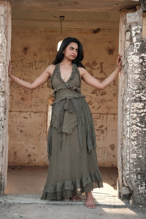 A woman wearing an olive green Rust Ember Nomad Maxi Dress with sleeveless, layered ruffled detailing standing in a stone doorway.