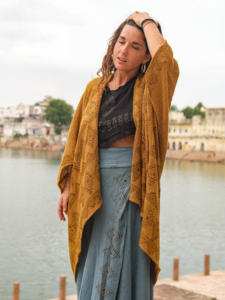 A woman with long brown hair stands by a riverside, eyes closed and hand to her head, wearing the Cocoon Shrug—hand block-printed in cotton—over a black top and blue skirt, with historic buildings in the background.