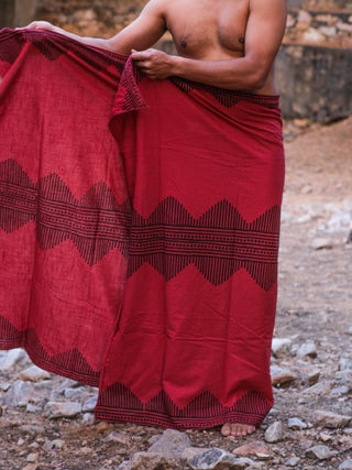 A person wears a red Arya Dhoti in traditional style, draped around the waist and standing barefoot on rocky ground with a bare upper body.