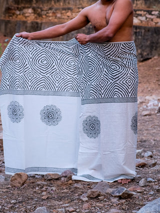Outdoors, a person displays the Sakshama Dhoti—a white traditional Indian garment featuring black geometric and circular designs—against a rustic, rocky backdrop that accentuates this elegant fashion piece.