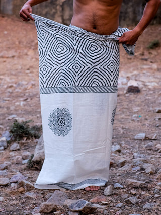 A person stands shirtless outdoors on rocky ground, wrapping a Sakshama Dhoti—a traditional Indian garment featuring black and white geometric and circular patterns—around their waist.