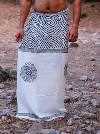 A person stands barefoot on rocky ground, wearing the Sakshama Dhoti—a white traditional Indian garment with black geometric and circular patterns—wrapped around the waist to the ankles, leaving the upper body uncovered.