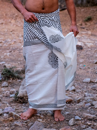 A person stands barefoot on rocky ground, adjusting a Sakshama Dhoti—an elegant white cotton dhoti with black geometric and circular patterns—at the waist. The upper body is partially out of view.