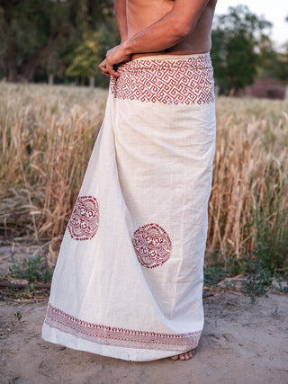 A person stands outdoors in front of a grassy field and trees, wearing the red Aastha Dhoti featuring traditional Rajasthani handblock print designs.