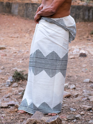 Wearing the Arya Dhoti in white with a black block print, a barefoot person stands on rocky ground, showing only their lower torso and legs.