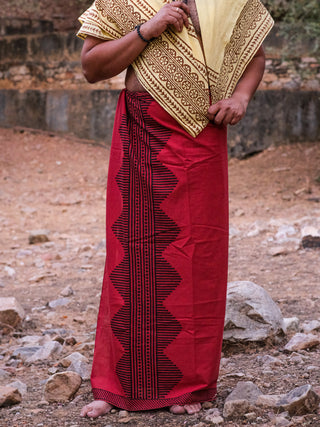 A person stands barefoot on rocky terrain, donning the Anviksha Dhoti with a red and black patterned sarong. Paired with a light brown block-printed shawl, their traditional Indian attire flows elegantly while concealing their face.