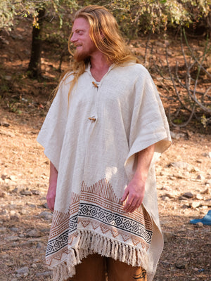 A person with long red hair stands outdoors, smiling and looking to the side while wearing a cream-colored poncho with geometric patterns and fringe, elegantly paired with the Swing Shawl draped over their shoulders.
