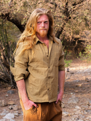A man with long blond hair and a beard stands outdoors in a natural, rustic setting of trees and rocks. He is wearing sandyellow YinYang Shirt and matching pants, the shirt’s subtle design adding an element of balance to his earthy ensemble.