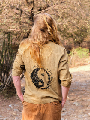 A person with long blond hair, wearing the YinYang Shirt—a tan cotton top with a bold black Yin-Yang symbol on the back—stands outdoors on a rocky path with dry trees in the background.