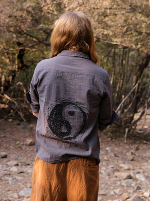 A person with long, light brown hair stands outdoors in a wooded area with rocks and trees, facing away. They are wearing the YinYang Shirt—a gray cotton shirt featuring a black yinyang symbol on the back—paired with light brown pants.