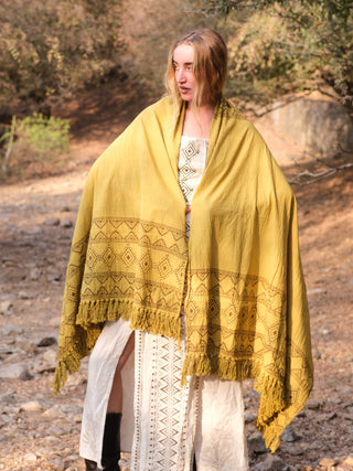 A person with long hair stands outdoors wrapped in a Neelkanth Shawl, its fringe and patterns reflecting bohemian style. They are in a long patterned dress and boots among trees and rocky terrain.