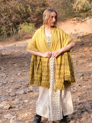 In a forest, someone with long hair is wrapped in a vibrant Neelkanth Shawl over a white dress with black geometric designs. The rocky ground, trees, and bushes complement this enchanting bohemian-inspired scene.