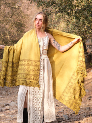 A person stands outdoors wearing a white dress with black patterns and a vibrant mustard yellow Neelkanth Shawl adorned with fringes. The shawl, featuring bohemian prints, is held open with outstretched arms against a backdrop of trees and rocks.