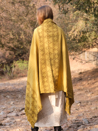 A light-haired individual stands on a rocky path wearing a Neelkanth Shawl adorned with intricate patterns, paired with a light dress and dark shoes, surrounded by lush trees, they embody bohemian charm.