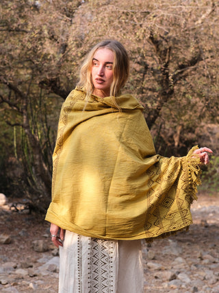 A person with long hair stands outdoors wearing a yellow Neelkanth Shawl with bohemian prints over a white outfit, amidst trees and rocky terrain, gazing to the side.