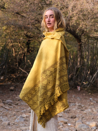 In a natural setting of dry trees and rocky ground, a person with long blonde hair outdoors wears a mustard yellow Neelkanth Shawl, featuring bohemian prints and fringe, amplifying its rustic charm.