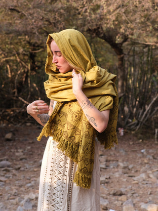 Wearing a light dress, a person stands outdoors on rocky terrain among trees, adjusting their mustard Neelkanth Shawl. Tattoos adorn their arms as they look down.