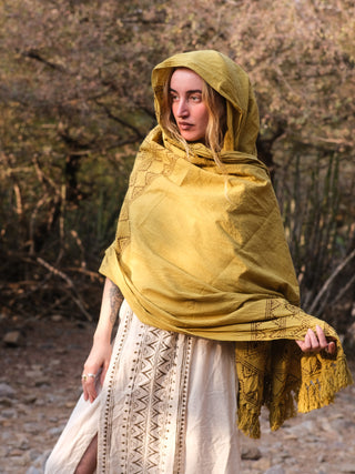 In soft daylight among trees and rocks, a woman with long blonde hair exudes elegance as she gazes to the side. She wears a vibrant Neelkanth Shawl with a hood over a patterned cream dress, showcasing subtle bohemian prints.