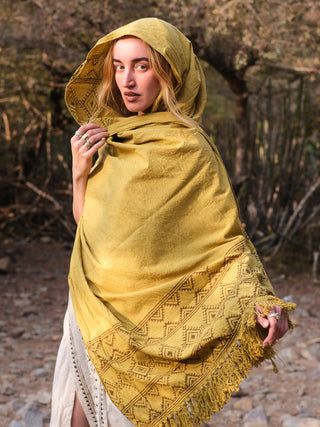 A person is outdoors in a woodland, draped in a Neelkanth Shawl with a hood and various silver rings. The rocky ground and tree-filled background highlight the elegance of their bohemian look.