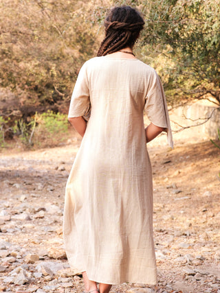 A person with long, braided hair stands on a rocky path, facing away from the camera, wearing the elegant Garga Dress with delicate block prints. Trees and dry foliage provide a serene outdoor backdrop.