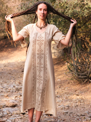 A woman with long dreadlocks stands outdoors on a rocky path, lifting her hair with both hands. She wears an elegant beige Garga Dress with block printing and a necklace, surrounded by trees and shrubs.