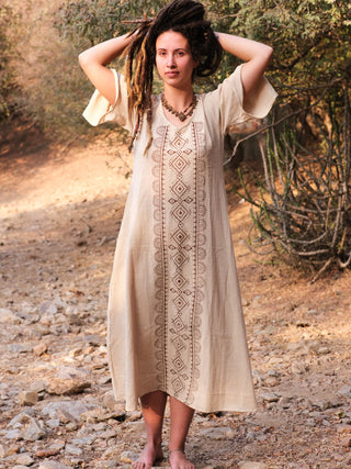 A person with long dreadlocks stands barefoot on a rocky path, surrounded by dry bushes and trees, wearing the Garga Dress with elegant geometric block printing.