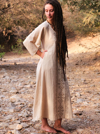 A barefoot woman with long brown dreadlocks stands on rocky ground, wearing the Garga Dress with detailed block printing. She gazes downward, surrounded by trees and bathed in warm sunlight.