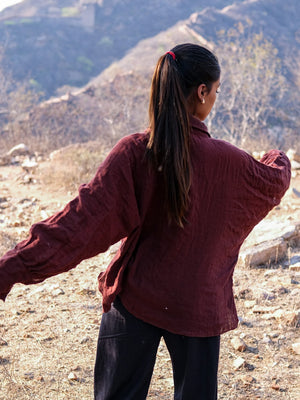 Standing outdoors with arms outstretched and facing away toward a rocky, hilly landscape, a person with long dark hair in a ponytail wears the loose maroon Ciraya Baggy Shirt paired with black pants.