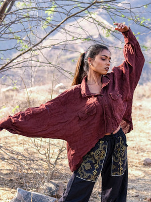 A woman poses outdoors in a loose maroon Ciraya Baggy Shirt and black pants with gold patterns, standing among branches and dry grass with one arm raised.