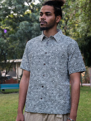 A man with a beard and tied-back hair stands outdoors wearing the Shipibo Shirt, which features a geometric black-and-white Shipibo print. Trees and a bench are visible in the background.