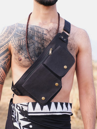 A person, with tattoos on their shoulder and chest, wears a Luca Festival Bag with two zippered compartments. Theyre adorned with a necklace and patterned wrap skirt, standing against a blurred natural backdrop.