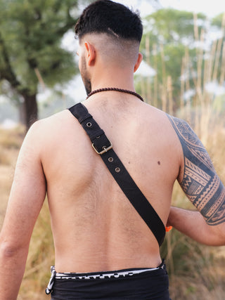 A shirtless individual stands outdoors with the Luca Festival Bag on their shoulder. They have a short haircut, a beaded necklace, and a tattoo on their left arm. Blurred trees and tall grass in the background highlight the festival vibe.