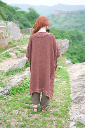 Back view of the model walking away in the brick red knit kimono, showing the plain knit back, the block printed collar and the cobweb trim along the hem, over olive harem pants on a stone path with green hills behind.