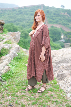 Full-length front view of the model in the brick red knit kimono with the pale block printed border, showing the deep side split, the mid-calf length and the cobweb fringe hem, worn over olive harem pants and sandals on a hillside above the Great Wall.
