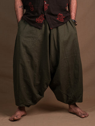 A person stands barefoot in green Sirwal pants, hands in pockets, wearing a dark shirt with red handprint designs. The loose-fitting pants and bold top add a modern twist to their look against a plain brown background.