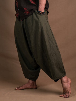 Barefoot on a tan surface, a person wears loose olive green Sirwal pants with their hand in the pocket and a black shirt with red patterns, showcasing only the lower half for a modern take on classic Sirwal pants style.