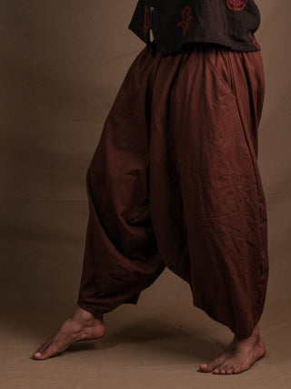 A person stands barefoot against a plain brown background, wearing loose Sirwal pants in brown with one leg bent and the other extended, giving this classic style a modern twist. The upper body is partially out of the frame.