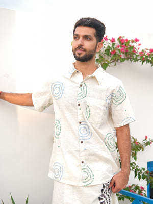 A man with a beard wears the Ishya Shirt—a short-sleeve, cream-colored premium fabric shirt featuring blue and green circular patterns—while standing beside a white wall with pink flowers and lush greenery in the background.