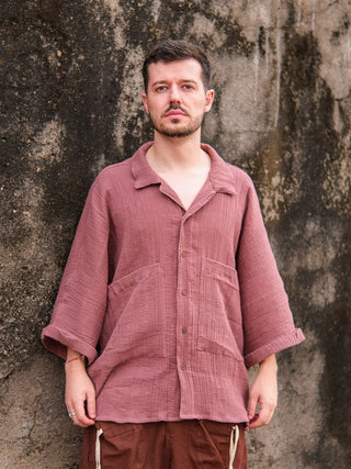 A man with short hair and a beard stands before a weathered wall, wearing the Yugma Baggy Shirt paired with brown pants for a relaxed, stylish look.