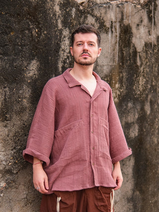 A man with short hair and a beard stands against a weathered wall, wearing the Yugma Baggy Shirt in mauve. Rolled sleeves and brown drawstring pants complement the shirts premium fabric as he gazes upward with a neutral expression.