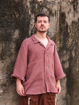 A man with short hair and a beard stands against a weathered wall, wearing the Yugma Baggy Shirt in mauve. Rolled sleeves and brown drawstring pants complement the shirts premium fabric as he gazes upward with a neutral expression.